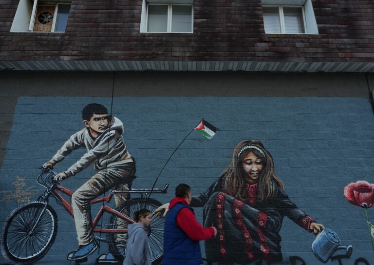 People are walking on the street in front of a large mural depicting a child on a bike with a Palenstinian flag