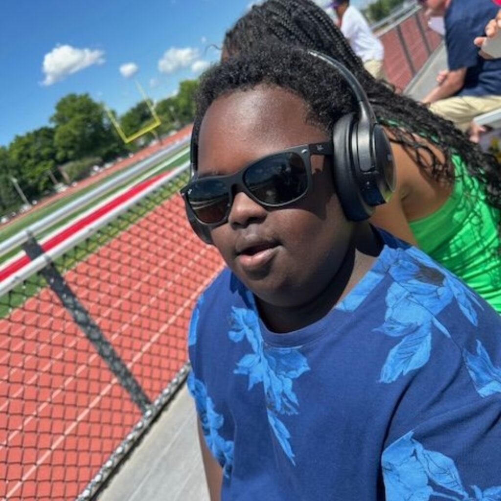 Bryan is sitting in the stands at the track, wearing sunglasses and headphones.
