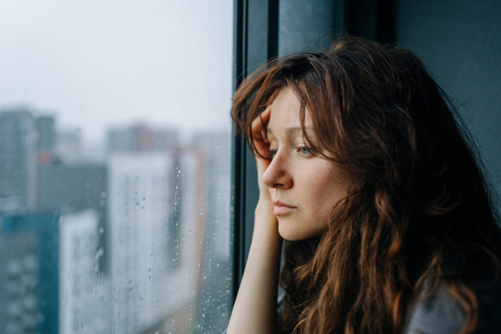 Young woman with brown hair stares through a rain-streaked window in a city apartment, pensive and melancholic, reflecting on loneliness and mental health during autumn A woman with brown hair looks sadly out of a window