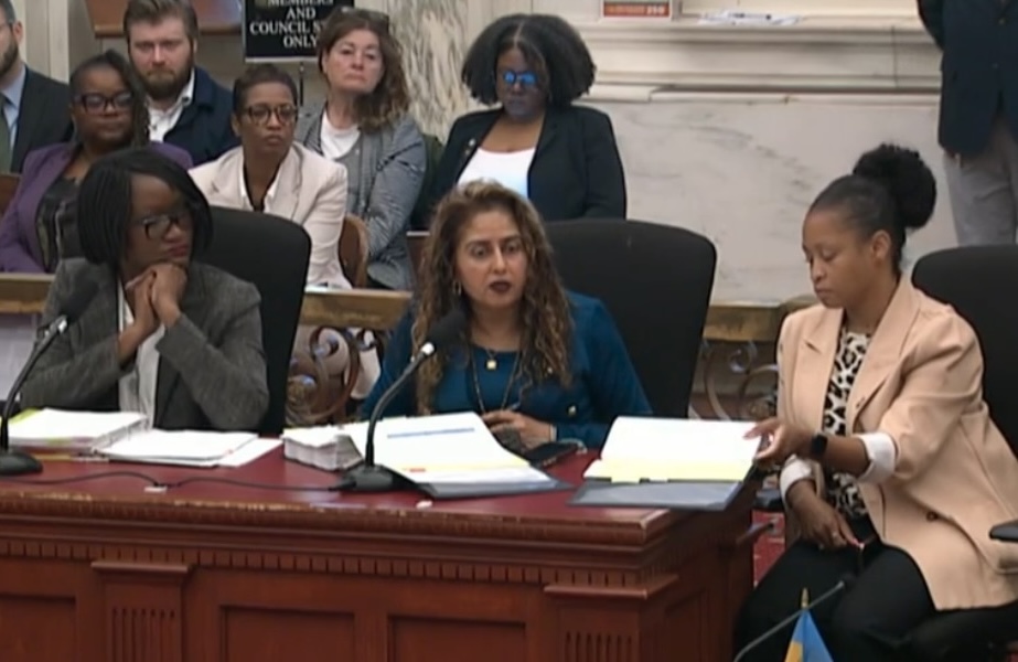 Three health officials are testifying at a large desk inside CIty Council chambers.