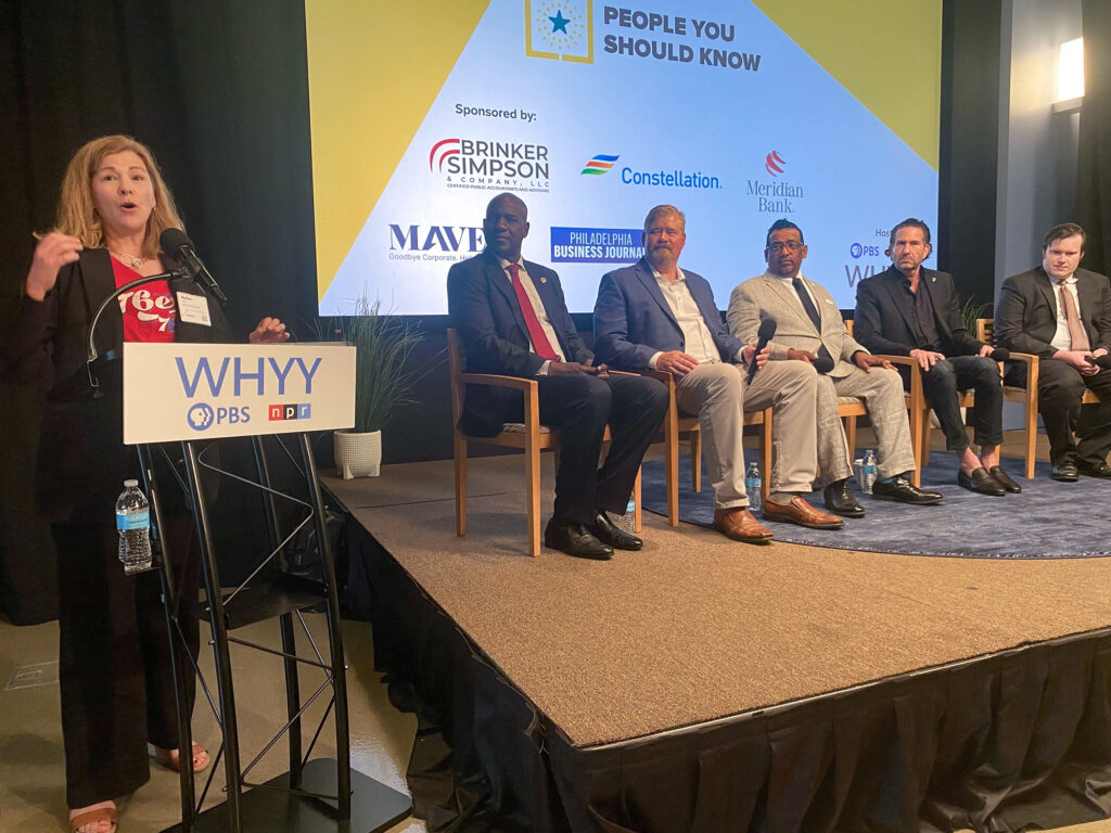 sports-economy-panel-j-mellman-2026-05-21 Maiken Scott is standing at a podium next to a stage which features five panelists sitting in a row.