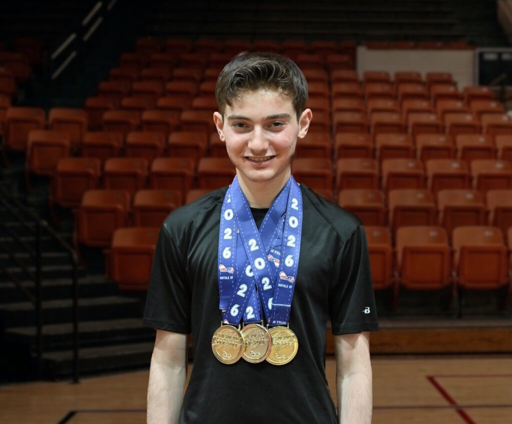 17-year-old Zach Peckman poses for a photo in a gym, and has 3 medals hanging around his neck.