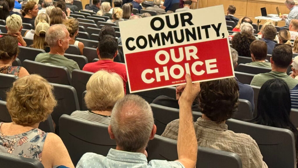 People at a town hall, one person holds a sign reading OUR COMMUNITY, OUR CHOICE