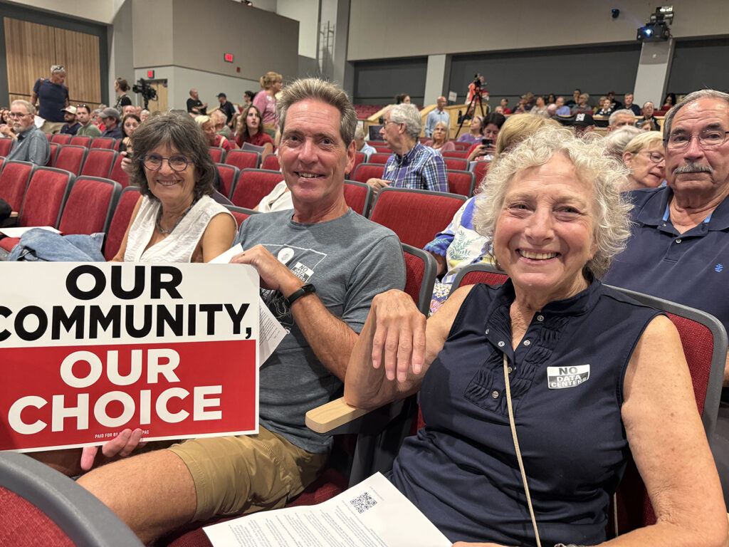 A man and a woman sit in an auditorium, with one holding a sign that reads "Our community, our choice"