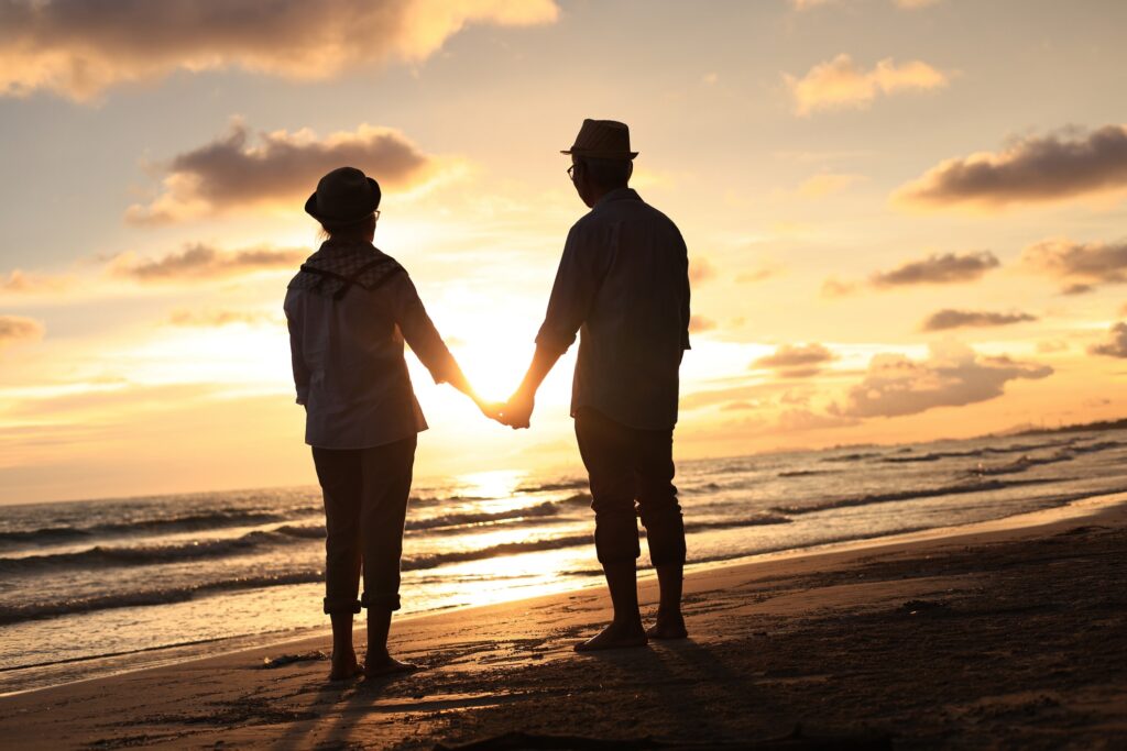 An elderly couple is walking on a beach at sunset holding hands.