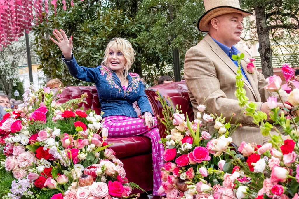 Obit Dolly Parton Dolly Parton waving to fans during a parade from a carriage, surrounded by pink and white flowers.