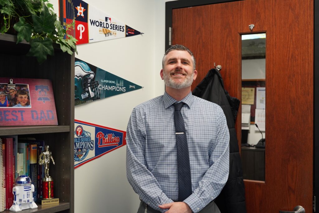Joe MacNamara is posing for a photo in his office at Radnor High School. On the wall are penants for the Phillies and Eagles