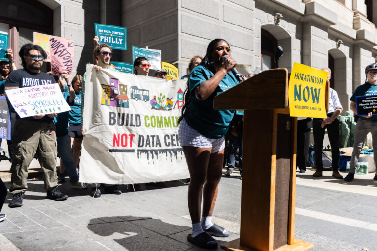 Sonya Sanders speaks at a podium with a crowd behind her holding anti-data center signs