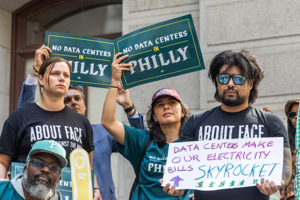 two people hold signs reading NO DATA CENTERS IN PHILLY, and another person holds a sign reading DATA CENTERS MAKE OUR ELECTRICITY BILLS SKYROCKET