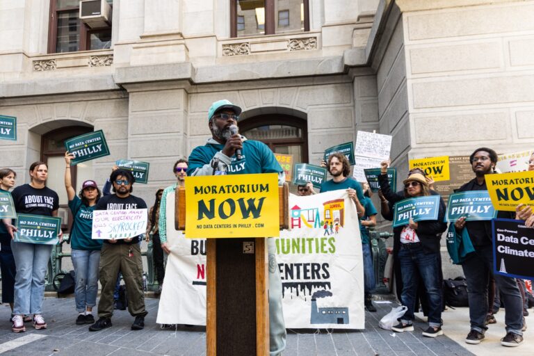 Shawmar Pitts speaks at a podium with a crowd behind her holding anti-data center signs