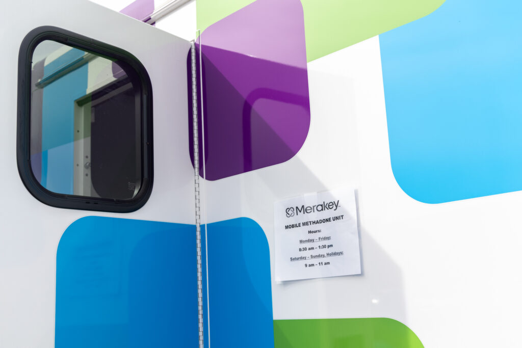 A printed sign is hanging on the side of the Mobile Methadone Unit displaying its hours of operation.