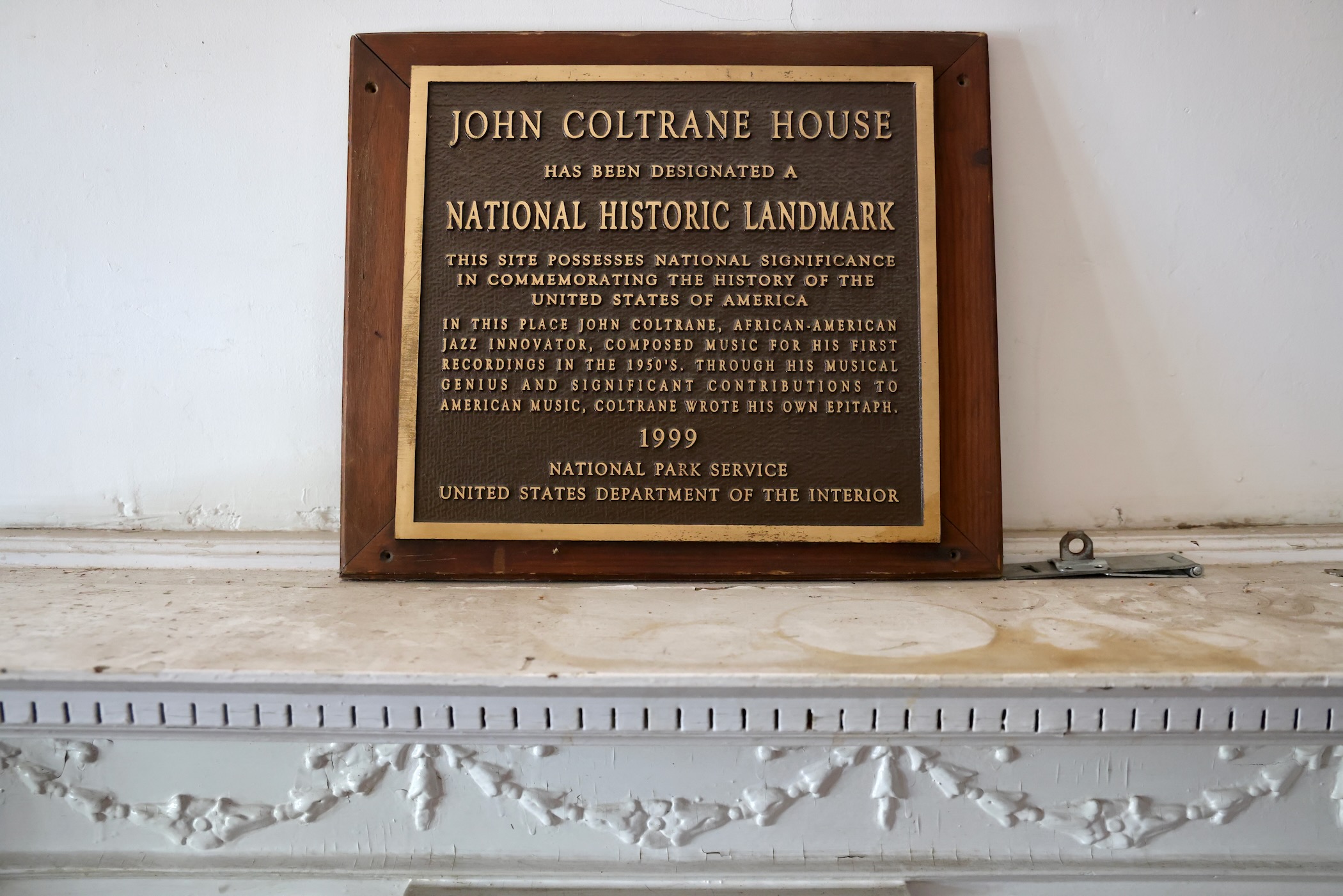 a plaque reads JOHN COLTRANE HOUSE HAS BEEN DESIGNATED A NATIONAL HISTORIC LANDMARK THIS SITE POSSESSES NATIONAL SIGNIFICANCE IN COMMEMORATING THE HISTORY OF THE UNITED STATES OF AMERICA IN THIS PLACE JOHN COLTRANE, AFRICAN-AMERICAN JAZZ INNOVATOR, COMPOSED MUSIC FOR HIS FIRST RECORDINGS IN THE 1950'S. THROUGH HIS MUSICAL GENIUS AND SIGNIFICANT CONTRIBUTIONS TO AMERICAN MUSIC, COLTRANE WROTE HIS OWN EPITAPH. 1999 NATIONAL PARK SERVICE UNITED STATES DEPARTMENT OF THE INTERIOR