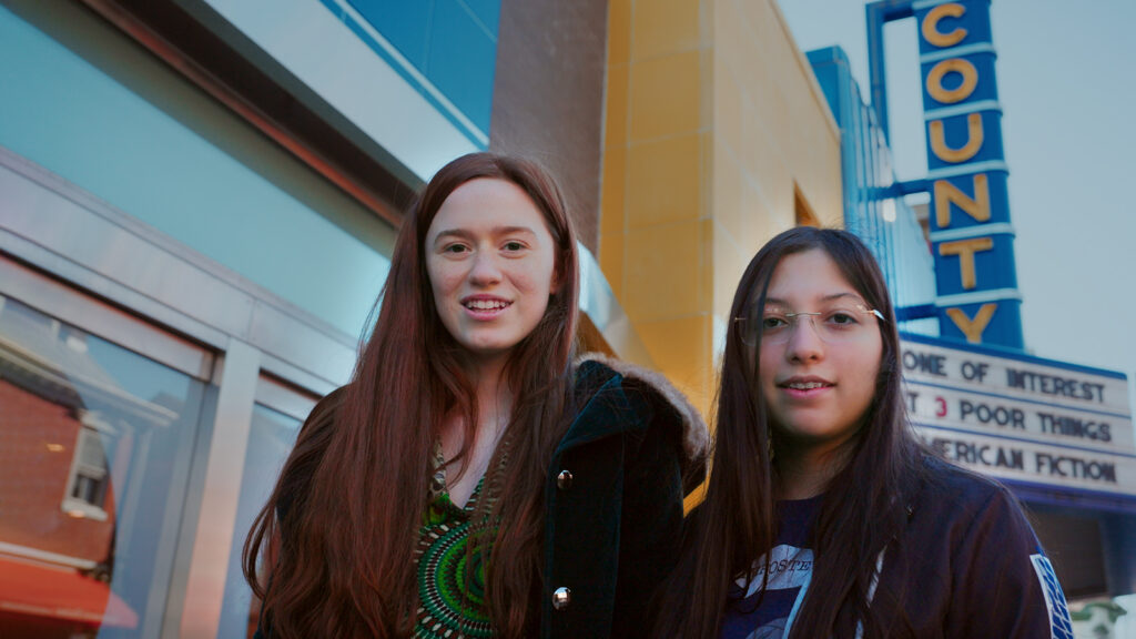 Two girls pose for a photo with the marquee of a movie theater behind them.
