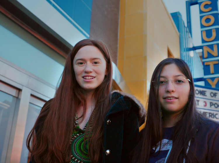 Two girls pose for a photo with the marquee of a movie theater behind them.