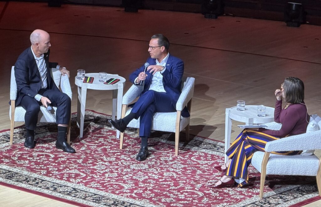 Cox-Shapiro-Garcia Josh Shapiro speaking into a microphone while sitting on stage at the Kimmel Center in white chairs with Spencer Cox and Lulu Garcia-Navarro.
