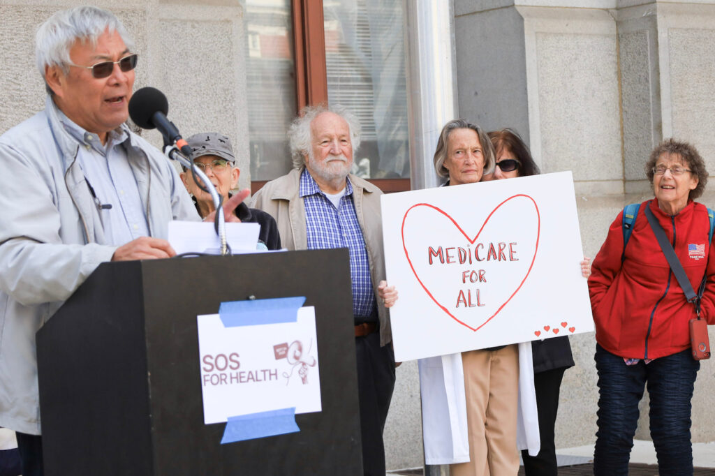 SOS rally-2026-philadelphia-medicaid-1-cropped A rallygoer speaks at a podium labeled SOS FOR HEALTH, and behind, a woman holds a sign reading MEDICARE FOR ALL in a heart