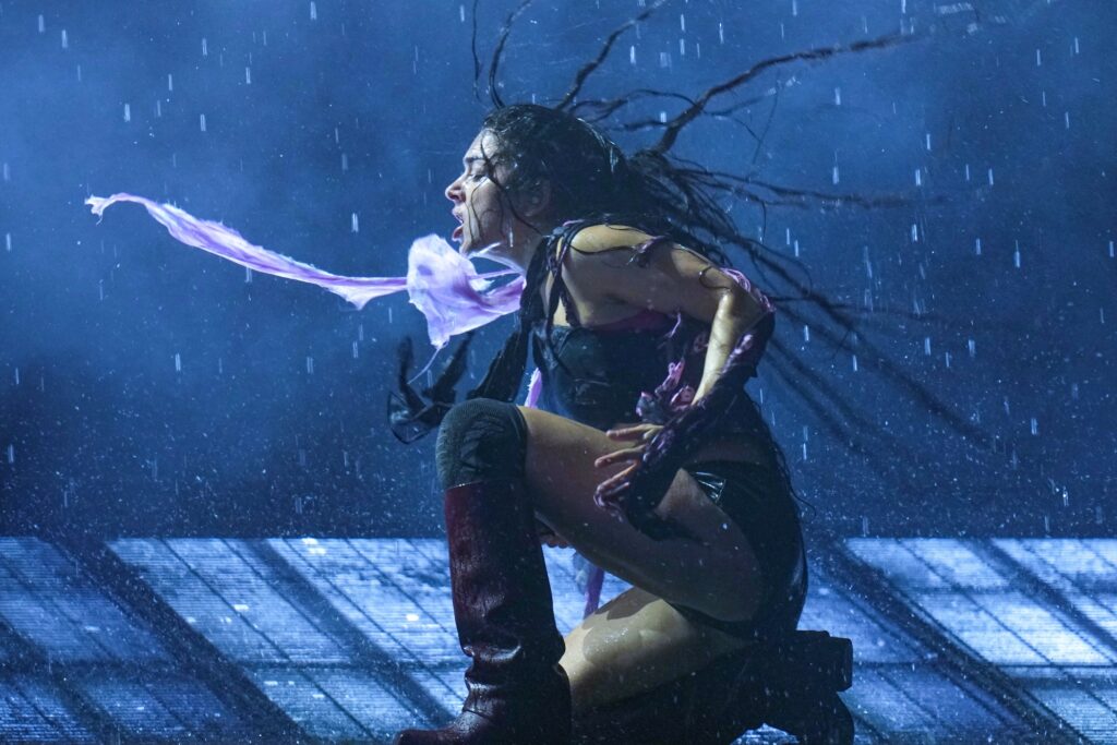 Charli xcx kneeling down with her purple scarf and hair flying in the air while performing on stage at Glastonbury.