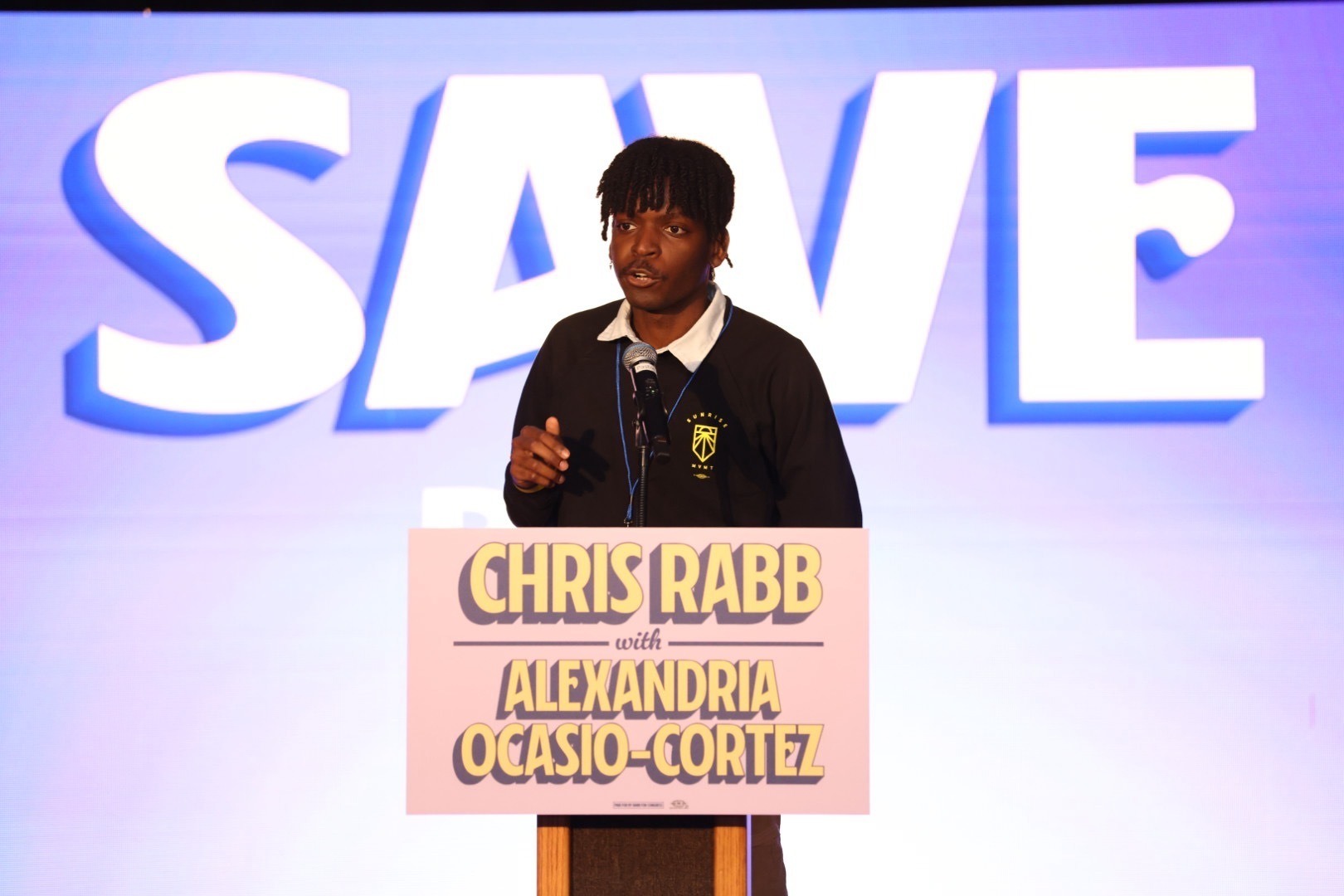 Sultan Smalley speaks at a podium reading CHRIS RABB WITH ALEXANDRIA OCASIO-CORTEZ in front of a backdrop reading WE SAVE US