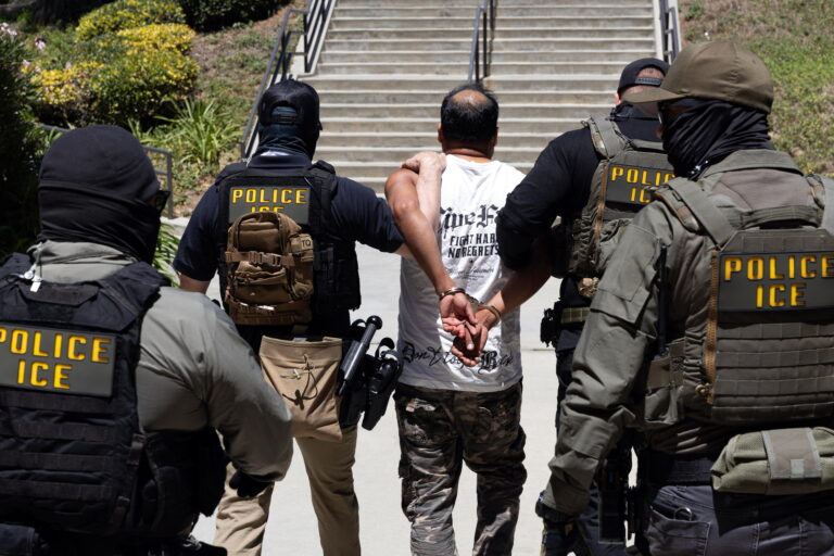 Four Immigration and Customs Enforcement agents walk a detainee out of the Ventura County Government Center in Ventura, California, on July 27. The individuals are photographed with their backs to the camera, and the detainee is handcuffed behind his back. The agents are wearing bulletproof vests that say 