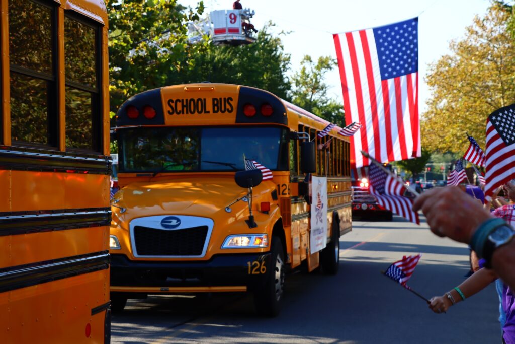School buses drive down the street as part of a parade, with people waving American flags.