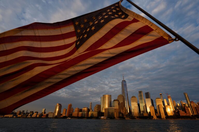 In the foreground, an American flag waves in the air. The skyline of lower Manhattan in NYC is in the background.