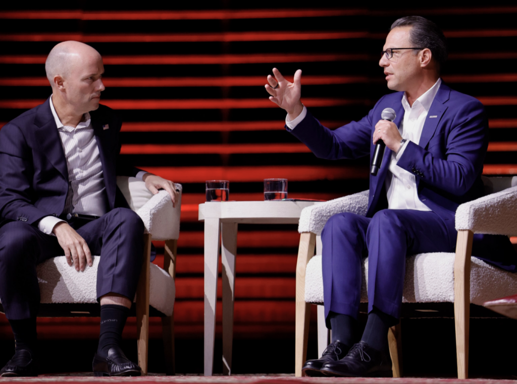 Josh Shapiro speaking into a microphone while sitting on stage in white chairs with Spencer Cox.