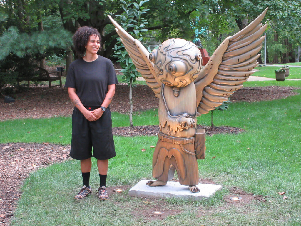 Visual artist Shantell Martin stands next to a 7-foot sculpture of an animal with wings, large eyeball and is bipedal