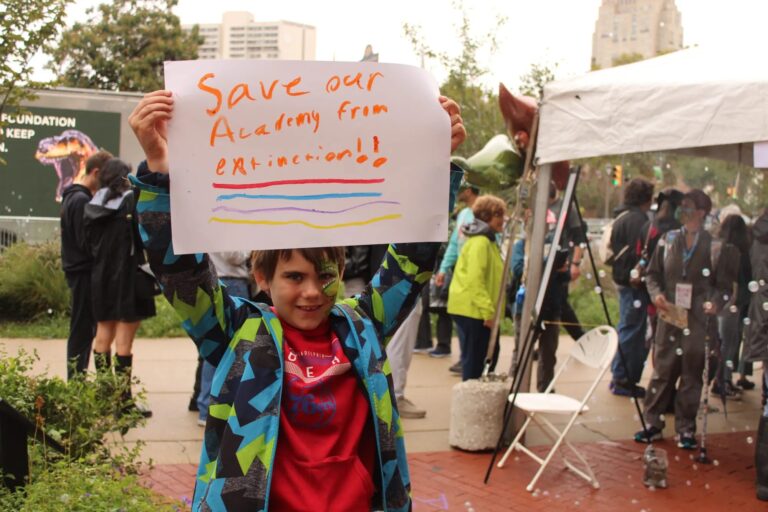 a child holds a sign reading Save our Academy from extinction!!