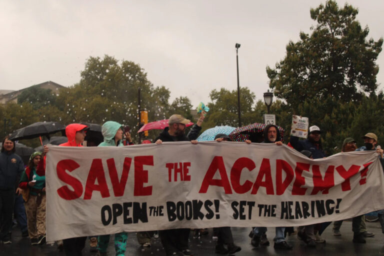 Marchers hold a banner reading SAVE THE ACADEMY! OPEN THE BOOKS! SET THE HEARING!