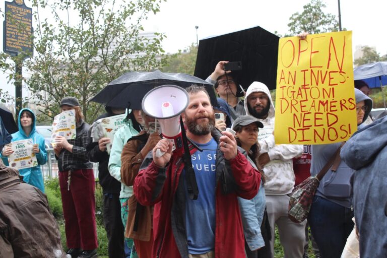 Corey Miller speaks in front of rallygoers, and a sign reads OPEN AN INVESTIGATION! DREAMERS NEED DINOS!