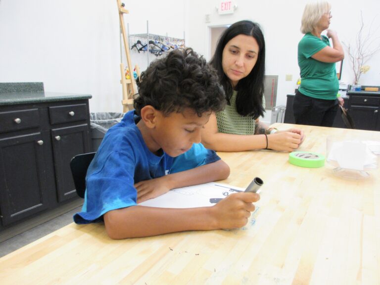 A child draws a picture at a table next to 2 adults
