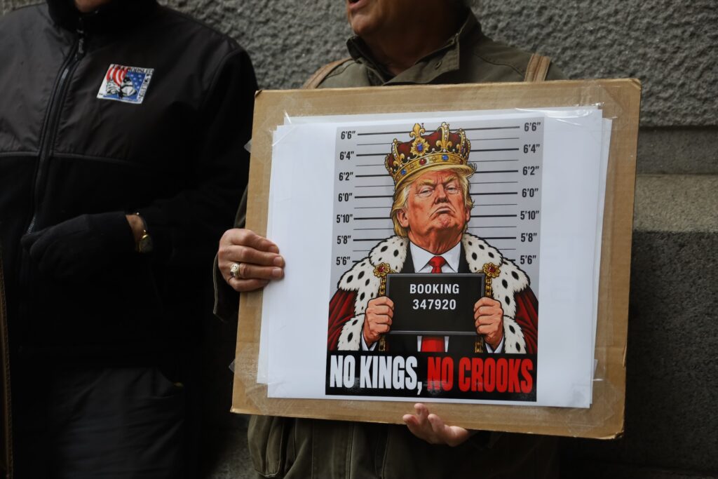 A protester holds up a sign depicting Trump wearing a crown and cape for a mugshot.