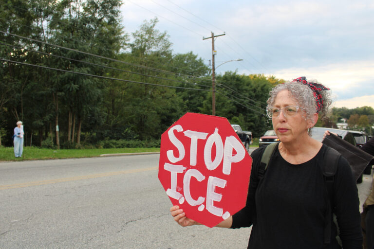 A woman is standing on the side of a road during an anti-ICE protest in Norristown, Pa. She holds up a handmade red stop sign that reads 'STOP I.C.E.'