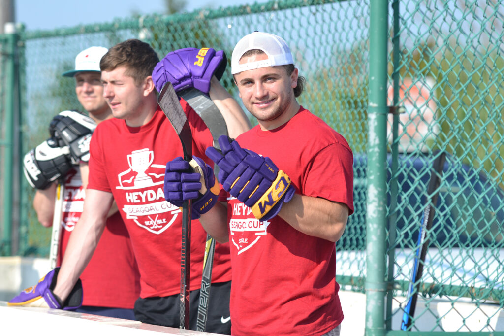 Men wait on the bench of an outdoor hockey rink holding their hockey sticks
